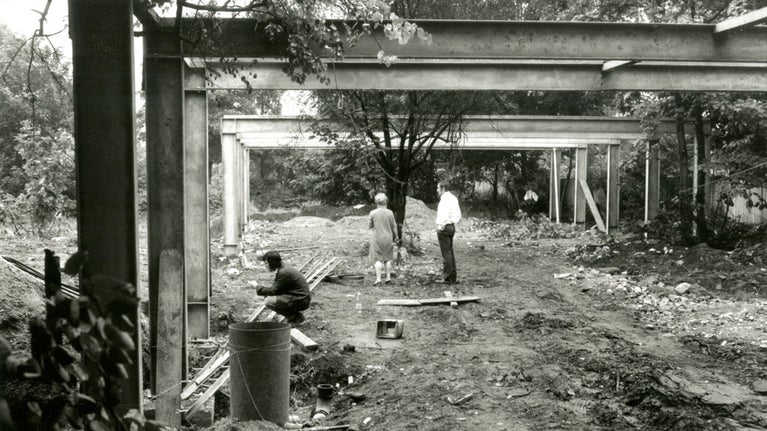 A black and white photo of a building site. Three people access the site. Steele beams form the skeletal shape of a single storey